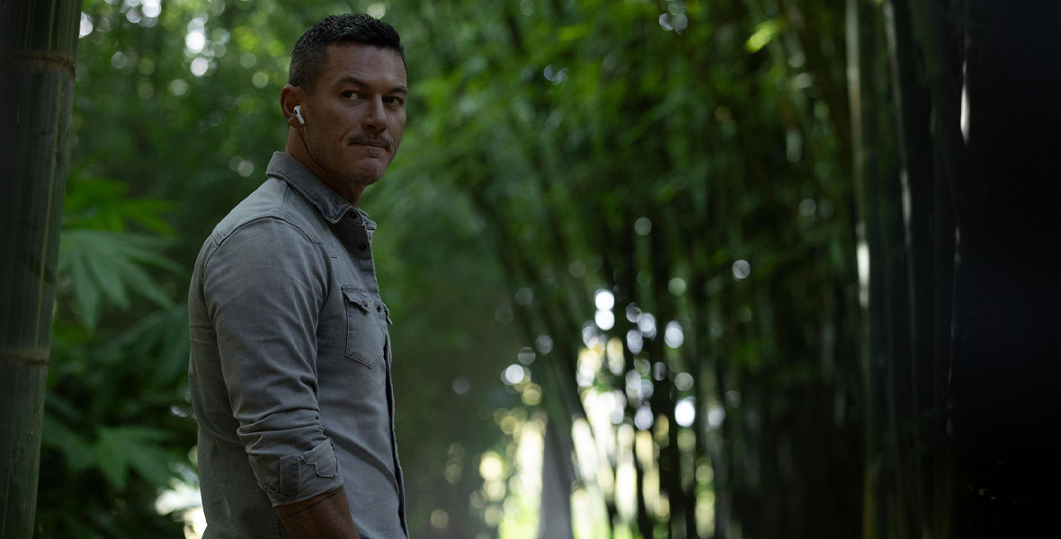 In a scene from Nine Perfect Strangers, Luke Evans stands in a forest with his hands in his pockets, wearing wired headphones and looking over his shoulder.
