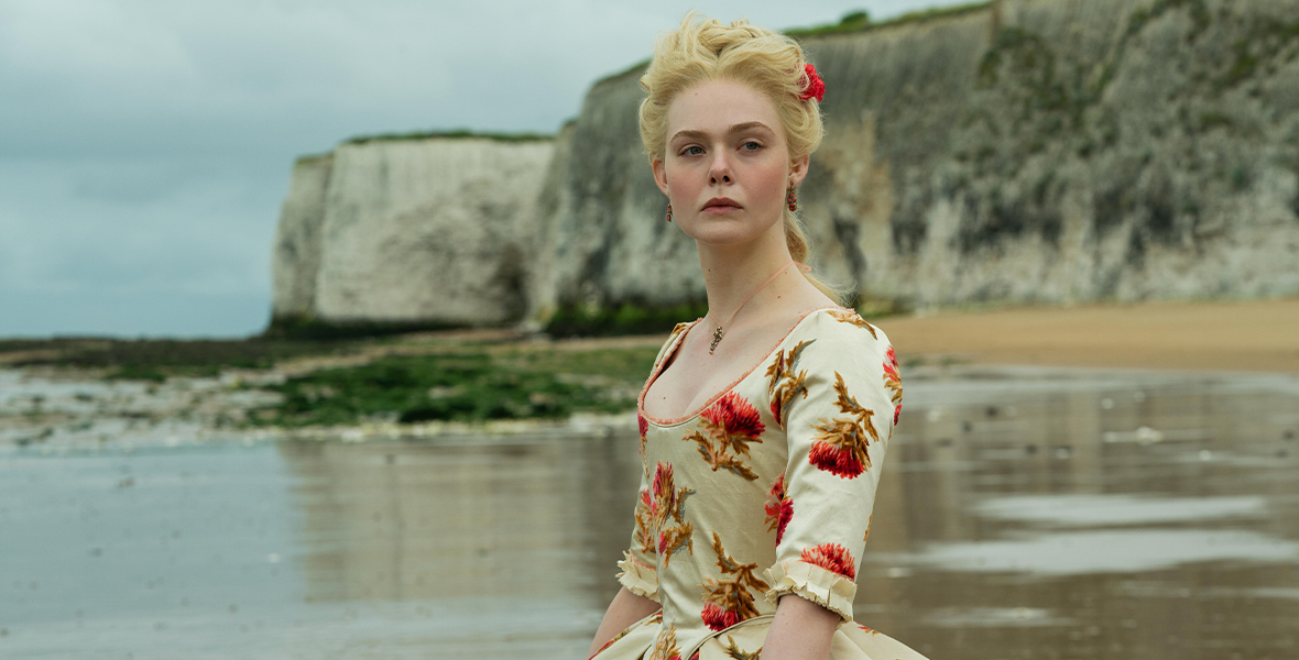 In a scene from The Great, Elle Fanning stands on the seashore, dressed in an elaborate gown covered in red flowers and staring off into the distance pensively.