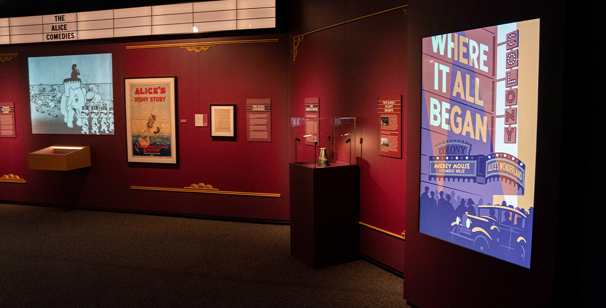 The entrance to the “Where It All Began” gallery in Disney100: The Exhibition. To the left is a display on The Alice Comedies, including a screen showing film clips and a poster for Alice’s Fishy Story against a red wall. To the right is the gallery poster, depicting a premiere at the Colony Theater for Steamboat Willie and Alice’s Wonderland.