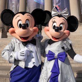 Mickey Mouse and Minnie Mouse, dressed in platinum outfits, stand together in front of The Franklin Institute in Philadelphia, Pennsylvania. Banners for Disney100: The Exhibition hang in front of the museum.