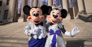 Mickey Mouse and Minnie Mouse, dressed in platinum outfits, stand together in front of The Franklin Institute in Philadelphia, Pennsylvania. Banners for Disney100: The Exhibition hang in front of the museum.