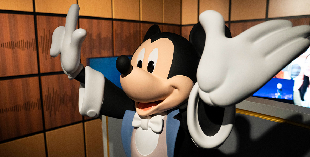 A statue of Mickey Mouse dressed as a conductor, with his hands raised, stands in front of tan and brown walls. The walls have a brown stripe of wood running across the middle that features the sound waves for the song “When You Wish Upon a Star.” 