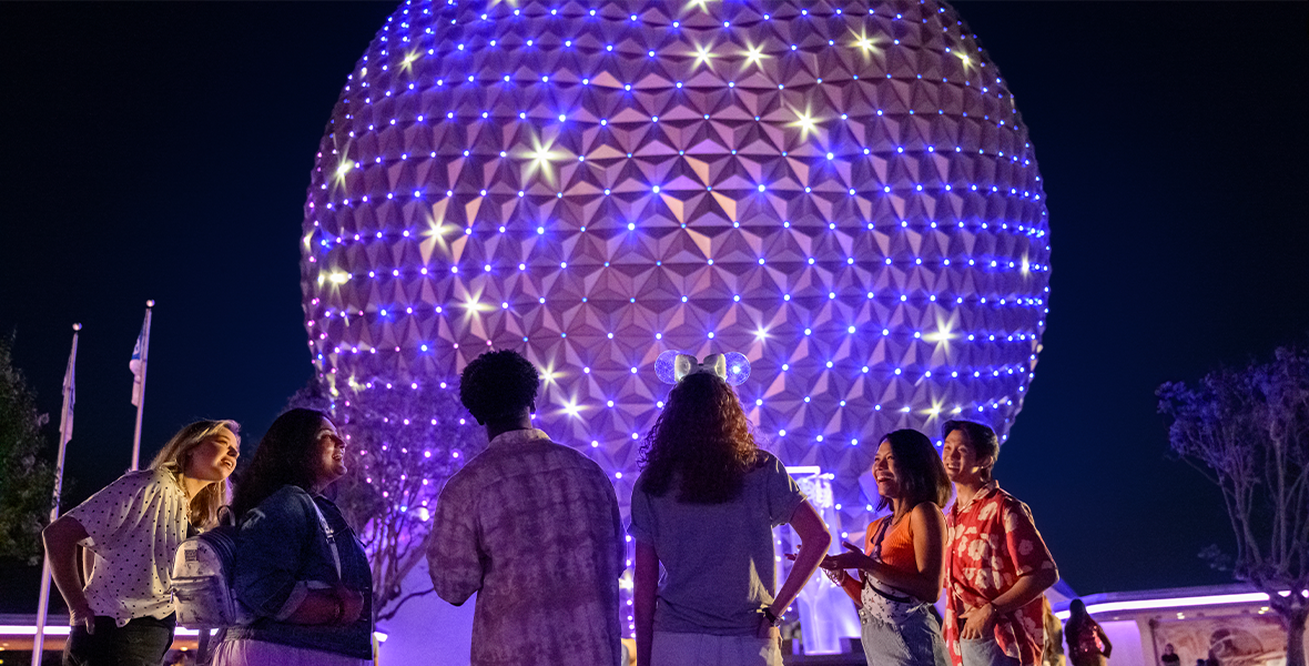Picture of EPCOT's Spaceship Earth, with it's glowing points of light shining in purple and white. Guests are standing in front of the iconic symbol of the park.
