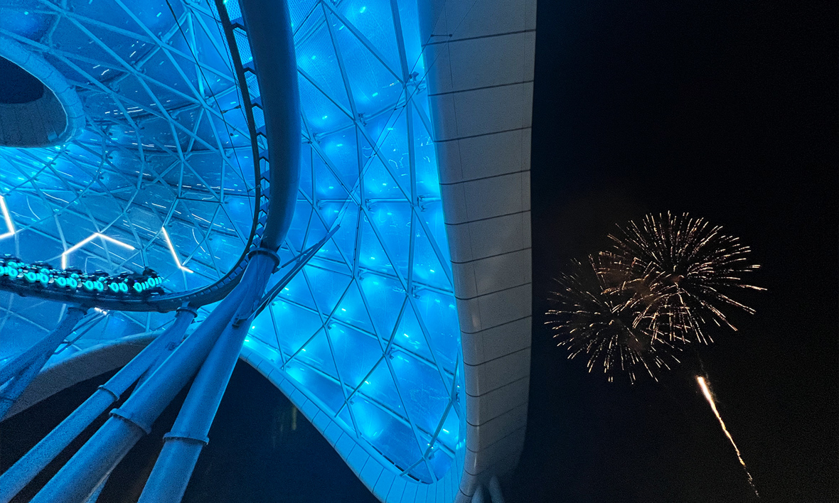 Lightcycles speed past on the TRON Lightcycle / Run attraction, as D23 Gold Members enjoy the cascading sparks of fireworks illuminating the sky, framed by the glowing canopy of the attraction.