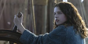 In Peter Pan & Wendy, 13-year-old Wendy grips the wheel of a ship and looks off into the distance. She wears a blue coat, and her brown hair flows freely just below her shoulders.