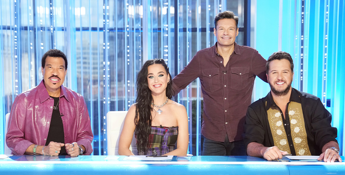 American Idol judges Lionel Richie, Katy Perry, and Luke Bryan sit at a large desk as host Ryan Seacrest stands behind them.