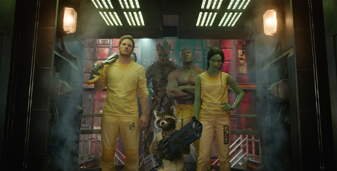 In a scene from Guardians of the Galaxy, actors Zoe Saldaña, Chris Pratt, and Dave Bautista stand in a doorway with Rocket, a raccoon, and Groot, a tree-like humanoid as they all wear yellow jumpsuits.