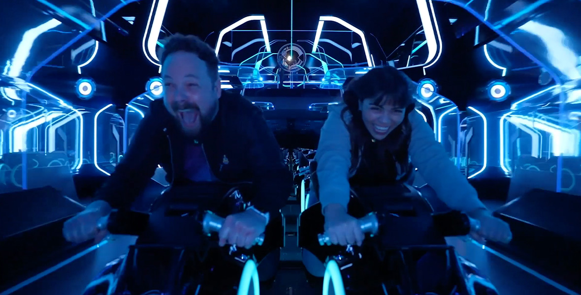 Mike Bithell and Heidy Vargas riding on TRON: Lightcycle / Run, both on the lightcycle-style attraction vehicles and wearing excited expressions.