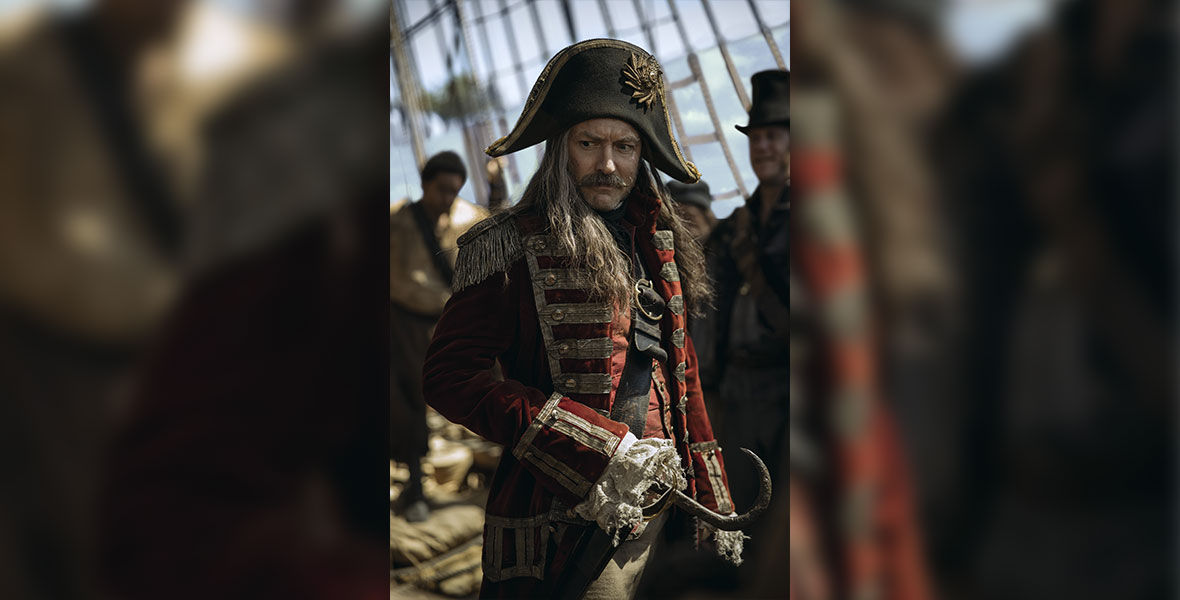 On his ship, Captain Hook stands and appears worried as his crew is blurry in the background. In place of his right hand is a rusting hook, and his graying hair is topped with a black pirate hat. He wears a red coat with gold adornments.