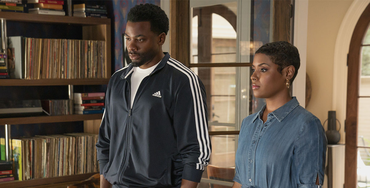In a scene from an episode of The Crossover, actors Derek Luke and Sabrina Revelle stand side by side inside a residential home. Luke wears a black tracksuit with three white stripes down the arms of the jacket and the side of the pants. Revelle wears a denim dress.