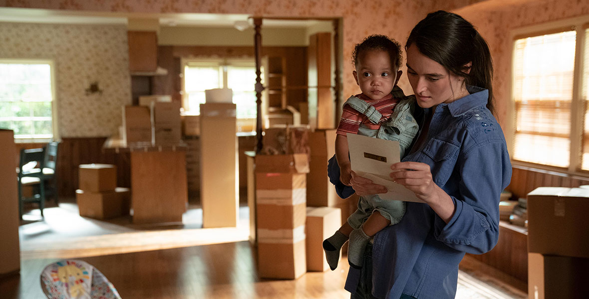 Young Clare, played by Sarah Pigeon, holds her baby daughter in one arm and a piece of paper in the other. Behind them are several unopened moving boxes.