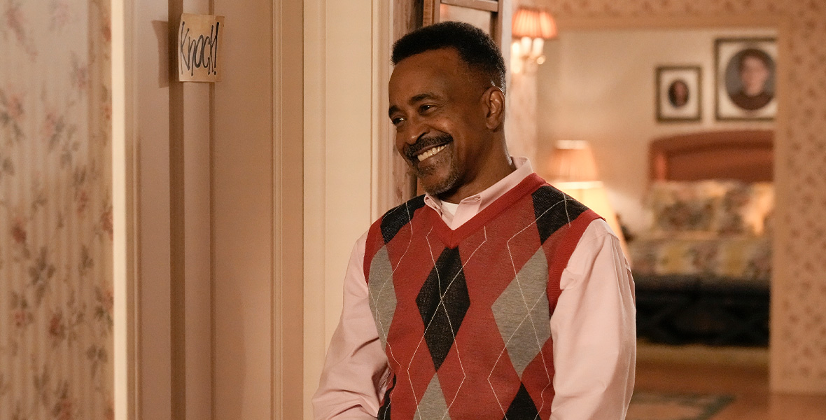 In a scene from an episode of The Goldbergs, actor Tim Meadows stands in a hallway and wears an orange and tan argyle sweater vest, tan pants, and a peach, button-down shirt.