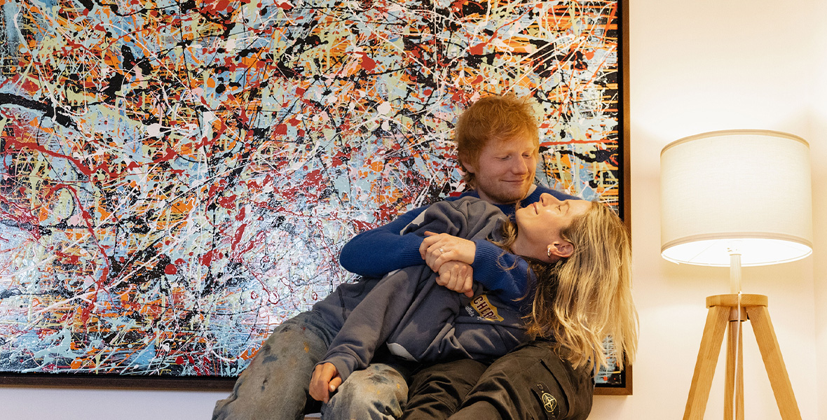 In a scene from Ed Sheeran: The Sum of It All, musician Ed Sheeran sits on a wooden cabinet and pulls his wife Cheryl in for a hug.