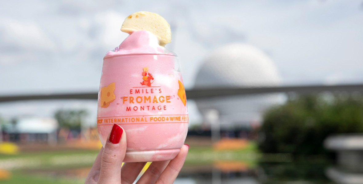 In an image promoting the 2023 EPCOT International Food & Wine Festival, a hand is holding a cup containing a yogurt-type treat with a cookie on top; the cup is labeled with the words “Emile’s Fromage Montage,” as well as images of cheese and Emile the rat from Ratatouille. EPCOT’s Spaceship Earth can be seen blurrily in the background.