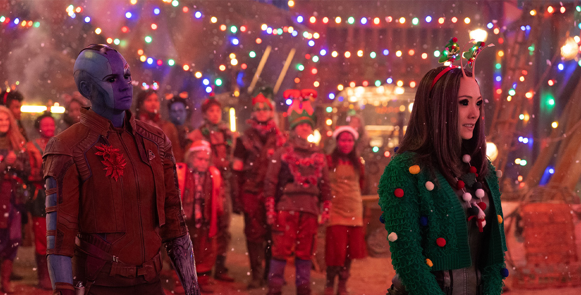 In a scene from The Guardians of the Galaxy Holiday Special, actors Karen Gillan and Pom Klementieff stand in front of a crowd. Klementieff wears a strand of colorful lights and a green, Christmas-themed sweater.