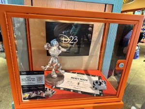 The D23 2023 Collector Set can be seen inside a glass case with bright orange trim, inside Island Mercantile at Disney’s Animal Kingdom Park. Against the back of the glass box is the D23 logo in platinum against a black background. Inside is the Mickey Mouse Leader of the Club statue standing on top of the D23 welcome certificate. Two membership cards can be seen, and they represent Disney Company characters and Marvel characters. On the left there is a QR code guests can scan to sign up for D23.