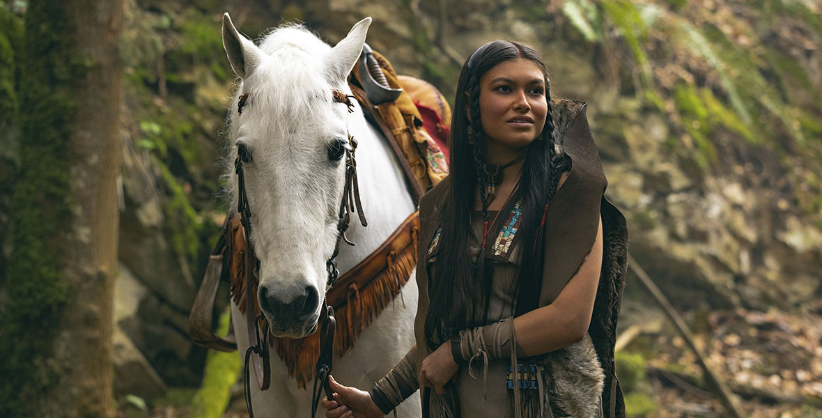 In the forest, Tiger Lily stands beside a white horse, guiding its reins as she looks off-screen. She wears Indigenous clothing and beading.