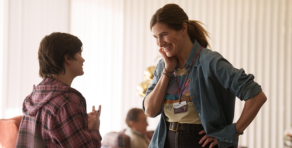 In a scene from an episode of Tiny Beautiful Things, actor Kathryn Hahn talks to actor Elizabeth Hinkler and wears a denim jacket and a yellow, graphic T-shirt.