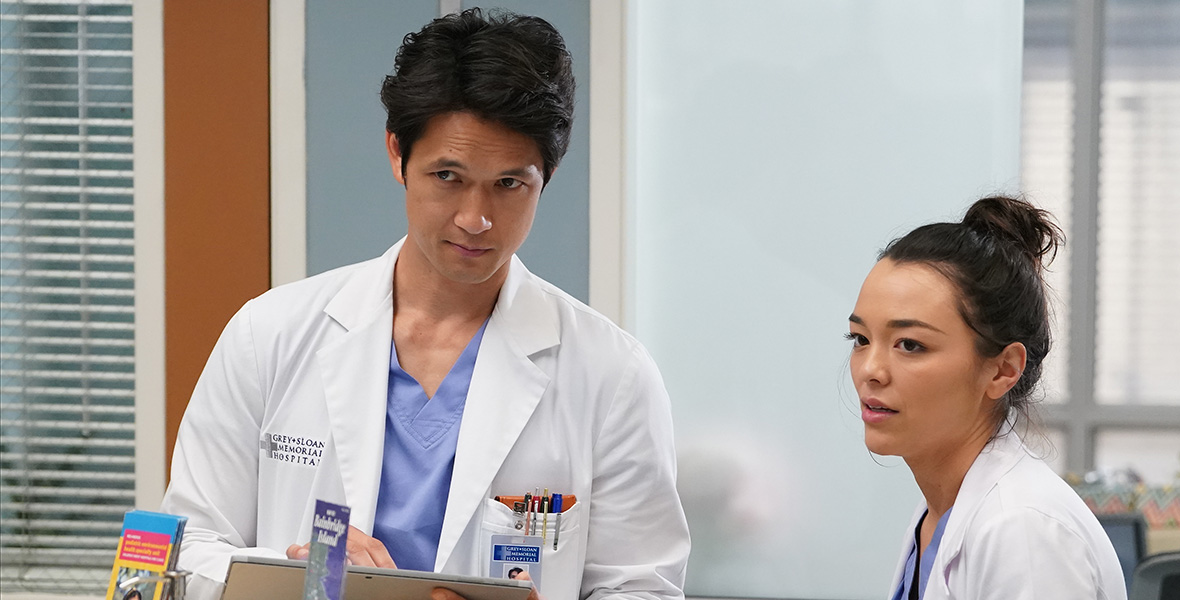 In a scene from Grey’s Anatomy, actors Harry Shum Jr. and Midori Francis stand in a hospital hallway, and both wear blue scrubs and a white lab coat.