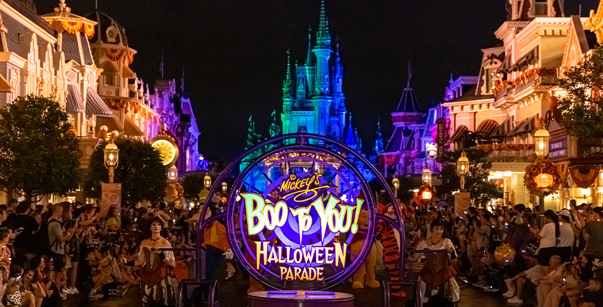 In an image promoting Mickey’s Not-So-Scary Halloween Party at Walt Disney World’s Magic Kingdom Park this fall, the front float with the logo for the Mickey’s Boo-To-You Halloween Parade is seen on Main Street, U.S.A.; it’s being steered by two performers on either side. Guests are sitting and standing on both sides of the street watching the parade, and all the Main Street buildings are lit up. Cinderella Castle can be seen in the background, silhouetted by the dark evening sky.