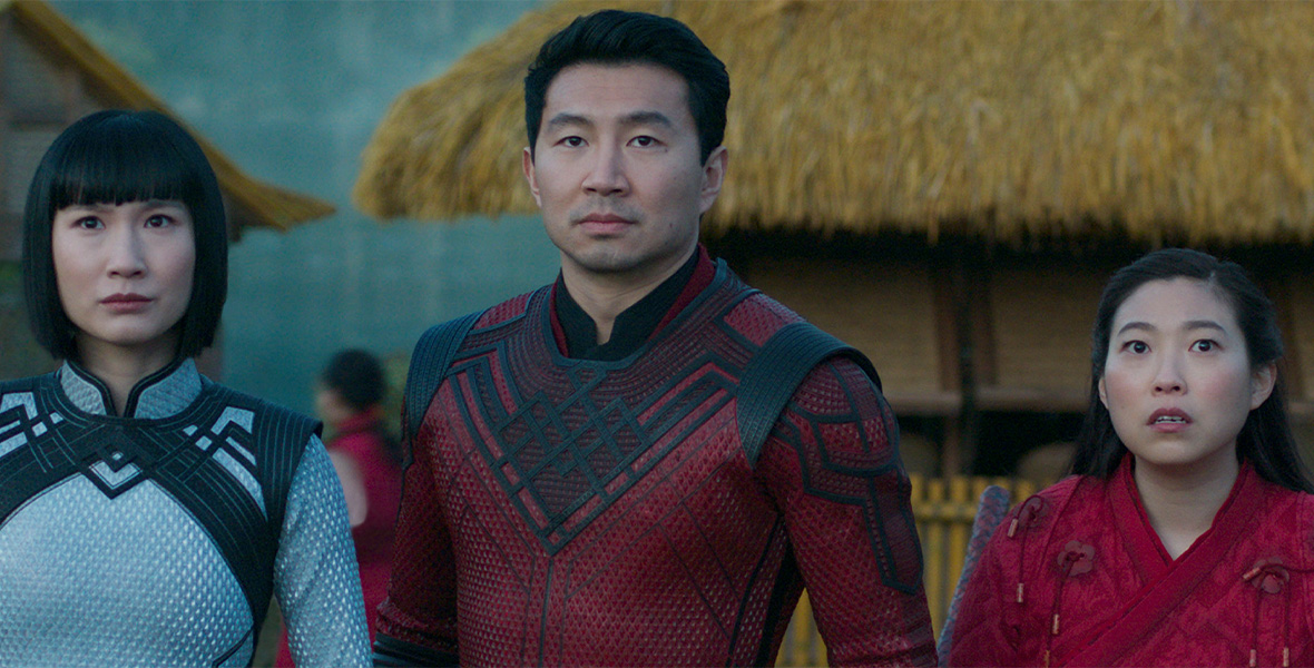 In a still from Marvel Studios’ Shang-Chi and The Legend of The Ten Rings, Xialing (Meng’er Zhang), Shang-Chi (Simu Liu), and Katy (Awkwafina) look concerned, off camera to the left. There are thatched huts behind them.