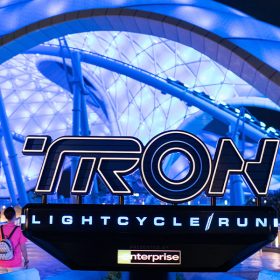 The entry plaza of TRON Lightcycle / Run presented by Enterprise as seen at night. The signage for the attraction is at center, with the dramatic illuminated canopy visible in the background. Groups of guests enter on the right, while another group exits at the left of the sign.