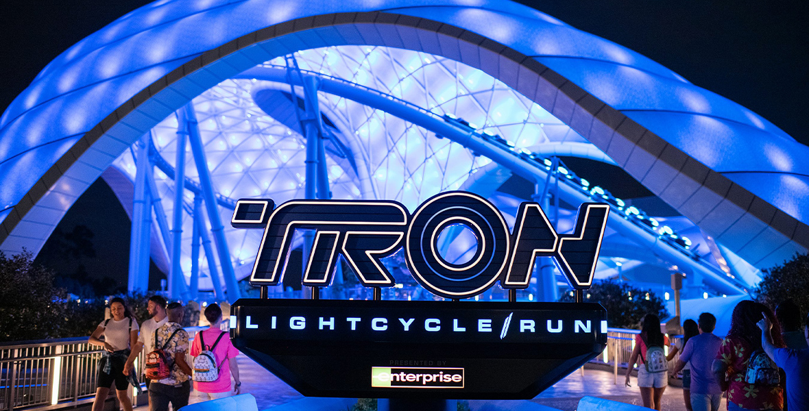 The entry plaza of TRON Lightcycle / Run presented by Enterprise as seen at night. The signage for the attraction is at center, with the dramatic illuminated canopy visible in the background. Groups of guests enter on the right, while another group exits at the left of the sign.