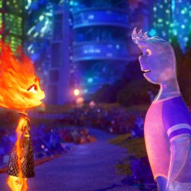 In a scene from the Pixar Animation Studios feature Elemental, Ember, a female fire person, stands facing Wade, a male water person, on a nighttime street with the colorful building of Element City behind them.