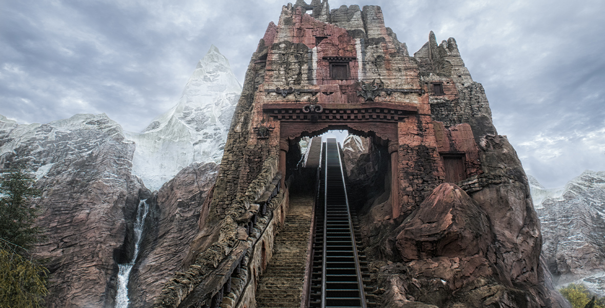 A view from the foot of the track of Expedition Everest – Legend of the Forbidden Mountain at Disney’s Animal Kingdom. It looks straight up at the ascending track, which goes through a crumbling monument and into the snowy peaks of Mount Everest. The cloudy sky blends with the frosty mountaintop.
