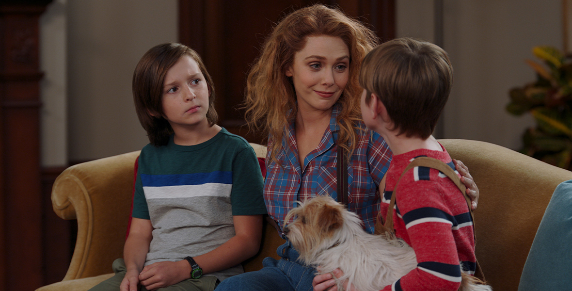 Wanda Maximoff, dressed and styled like a ’90s sitcom mother, puts her arms around her children, Tommy and Billy. Wanda is sitting in between both boys on a yellow couch, and Billy is looking directly at her with his dog, Sparky, in his arms.