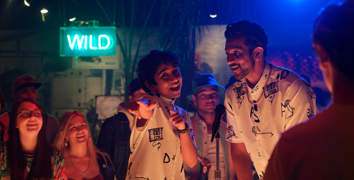 In a first-look image from Disney+’s World’s Best, Prem (Manny Magnus) is standing in front of a mic, next to his father (Utkarsh Ambudkar), performing for a small crowd. They’re wearing matching black and white shirts, and both are smiling. The crowd around them is smiling back at them. There’s a blue neon sign in the background that reads “WILD.”