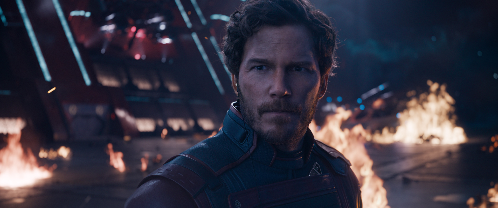 An image of Chris Pratt as Star-Lord in Guardians of the Galaxy Vol. 3. He stares ahead with a serious expression as areas of the spaceship he stands in catch fire.