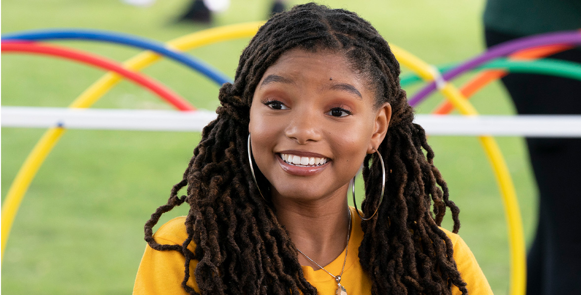 In the series Grown-ish, Halle Bailey plays college student Sky. Sky sits at an outdoor table with some chips and smiles brightly. She wears a yellow T-shirt, hoop earrings, and a necklace.