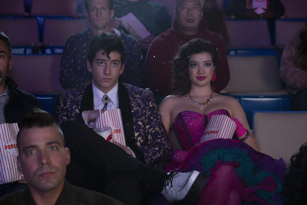 Milo Manheim and Peyton Elizabeth Lee sit side by side in a dark movie theater. They are both dressed in 80’s style formal attire. Milo has on a blue suit jacket patterned with gold detail. His hair is styled in an 80’s jheri curl and he has a silver bolo around his neck. Elizabeth is wearing a bedazzled pink sweetheart-neck dress, matching pink gloves, and costume jewelry accessories. They are both staring ahead at the screen in front of them and eating popcorn.