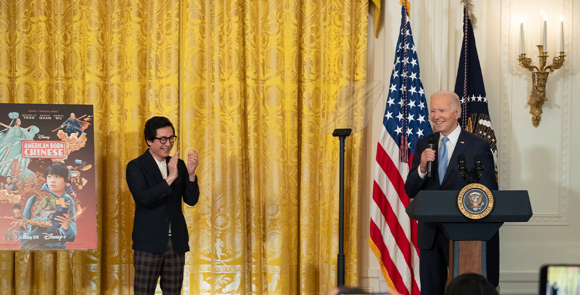 Actor Ke Huy Quan and stands on a small stage at the White House, to the left of President Joe Biden, who is at a lectern holding a microphone; they are both smiling. Quan is clapping; to his left is a poster for Disney+’s American Born Chinese, and behind him is a golden curtain.