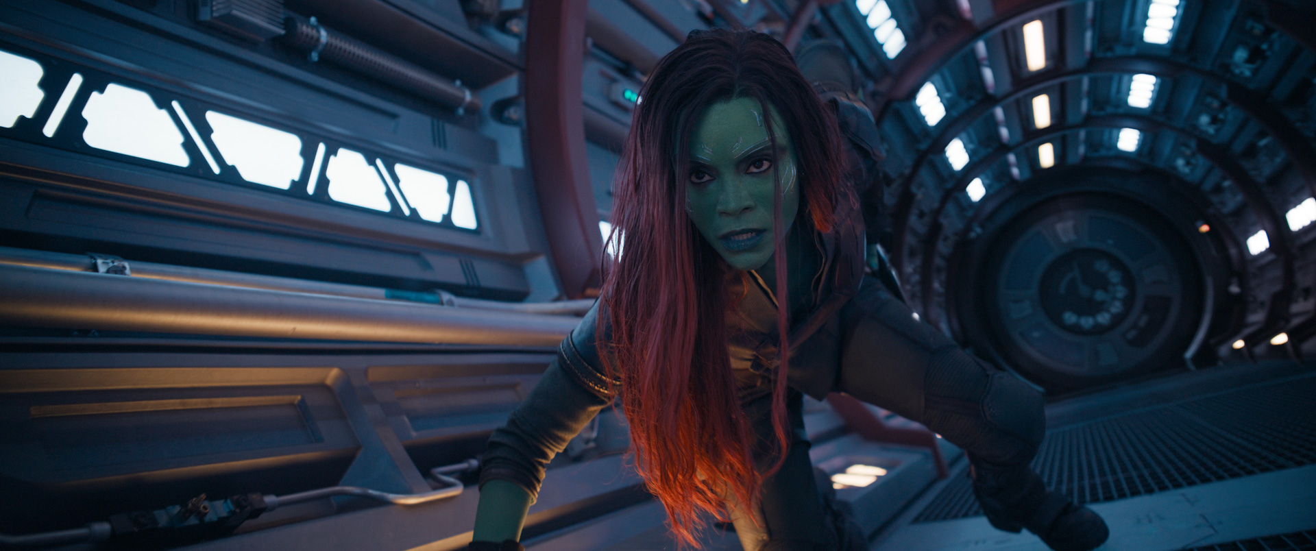 An image of Zoe Saldaña as the green alien Gamora in Guardians of the Galaxy Vol. 3. In the halls of a spaceship, she kneels on the ground and looks ahead. Her left arm reaches for a weapon holstered at her hip.
