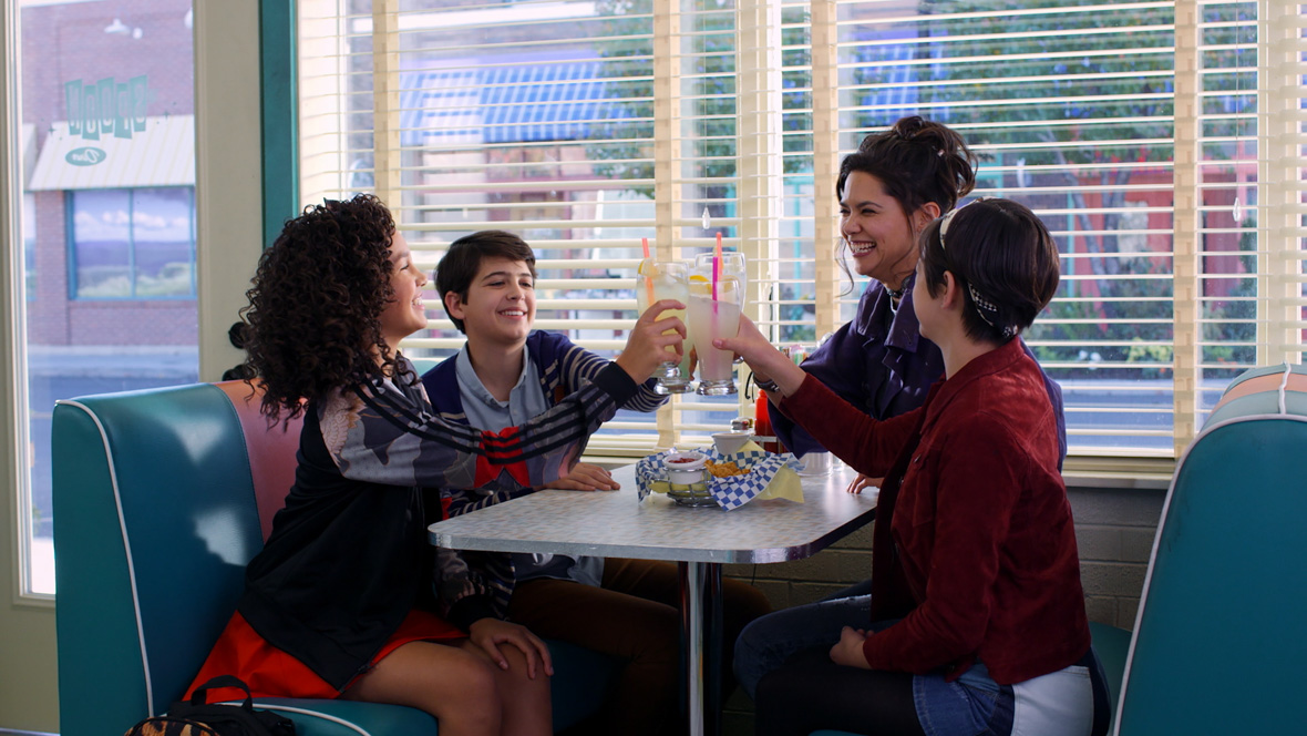 A screenshot from Andi Mack featuring Joshua Rush, Sofia Wylie, and Peyton Elizabeth Lee cheering their lemonades at a diner. Most of the cast is facing inward at each other, obscuring their faces, but Joshua can be seen clearly, smiling as he holds up his drink. The setting looks like a classic 50’s style diner with leather seats. Behind the cast you can see a suburban downtown street out of the restaurant’s window.