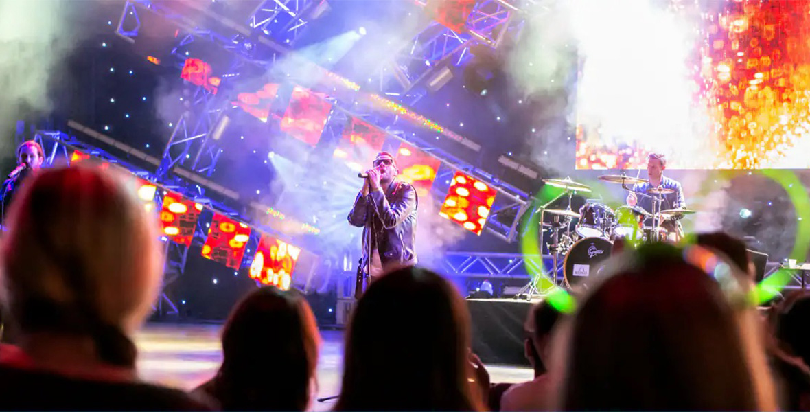 In a promotional image for the Eat to the Beat Concert Series at Walt Disney World Resort, a rock band is performing on a smoky stage, complete with a lead singer, a guitarist, and a drummer. Cool lighting is seen all around them. In the foreground, audience members are looking up at the stage.