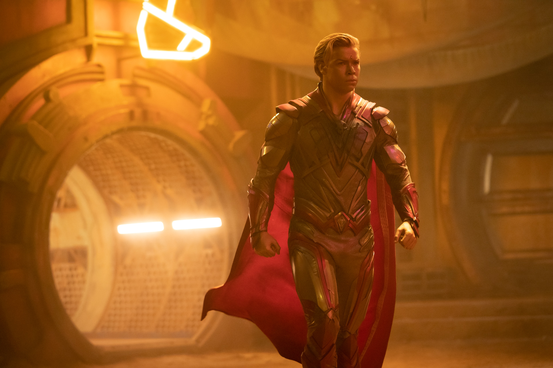 Will Poulter stands tall as Adam Warlock in Guardians of the Galaxy Vol. 3. The room he stands in glows gold, while Will wears a metallic suit and red cape. His blond hair is combed to one side.