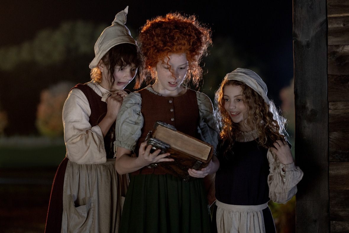 A scene from Hocus Pocus 2 featuring Taylor Henderson, Nina Kitchen, and Juju Brener as the young Sanderson Sisters. The three girls stand in the dark outside, peering down at Winifred’s iconic one-eyed spellbook. The trio are dressed in traditional 17th century costumes and are looking at the book in Taylor’s hands with worried and excited expressions.