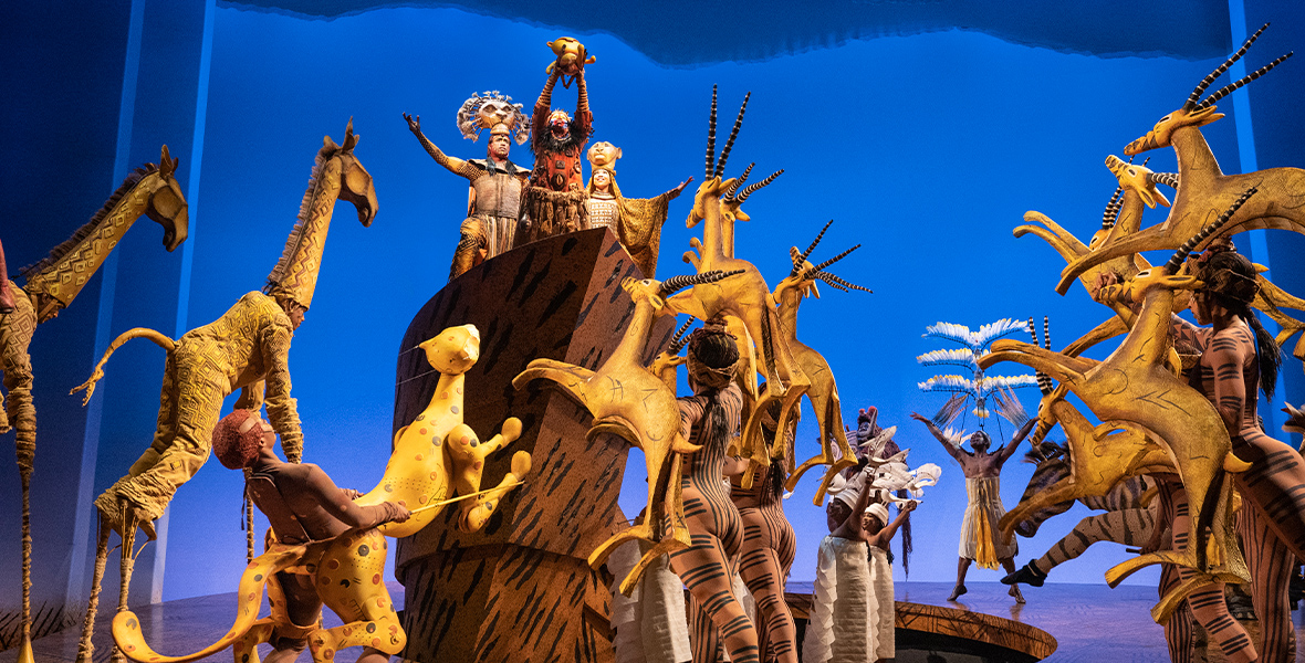 A production still from Disney’s The Lion King on Broadway, from the number “Circle of Life.” Puppet and human-hybrid representations of animals are seen across the stage, facing upstage and looking up at a set piece denoting Pride Rock. On said set piece, from left to right, Mufasa, Rafiki, and Sarabi are looking up at baby Simba (a puppet), being held in Rafiki’s hands. The stage background is a deep, bright blue.