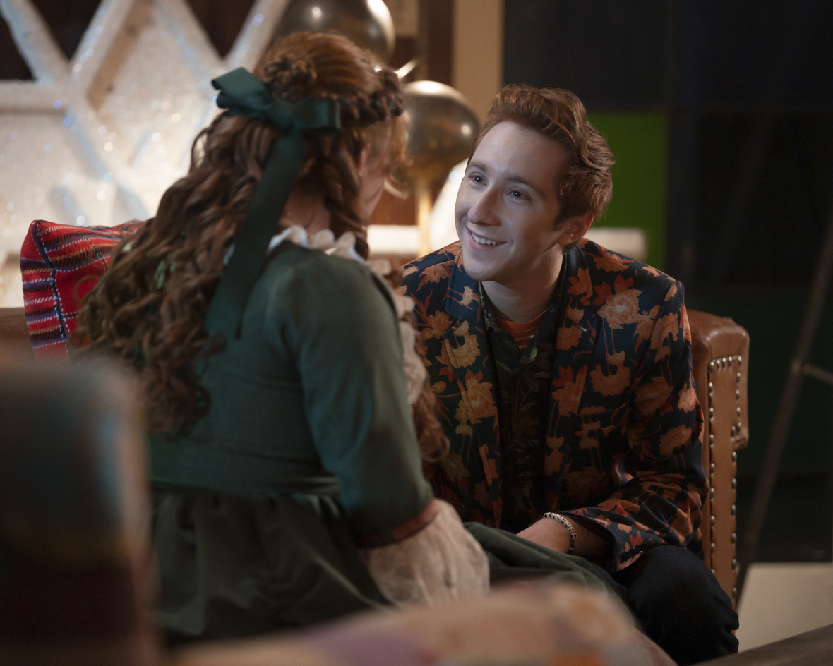 Larry Saperstein sits on a leather chair in front of Julia Lester, smiling up at her with adoration in his eyes. He is wearing a blue suit jacket with golden and brown autumn leaves all over it. A friendship bracelet can be seen on his left wrist and his red hair is styled casually. You can see Julia Lester from the back, and she is wearing her Belle costume from the Beauty and the Beast musical they are performing in High School Musical: The Musical: The Series.