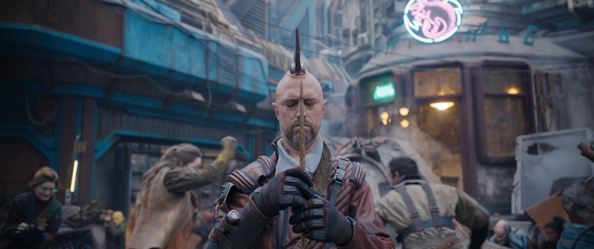 An image of Sean Gunn as Kraglin in Guardians of the Galaxy Vol. 3. Channeling his metallic mohawk, he shuts his eyes and grips the Yaka arrow to concentrate. Around him, people are running through a chaotic marketplace.