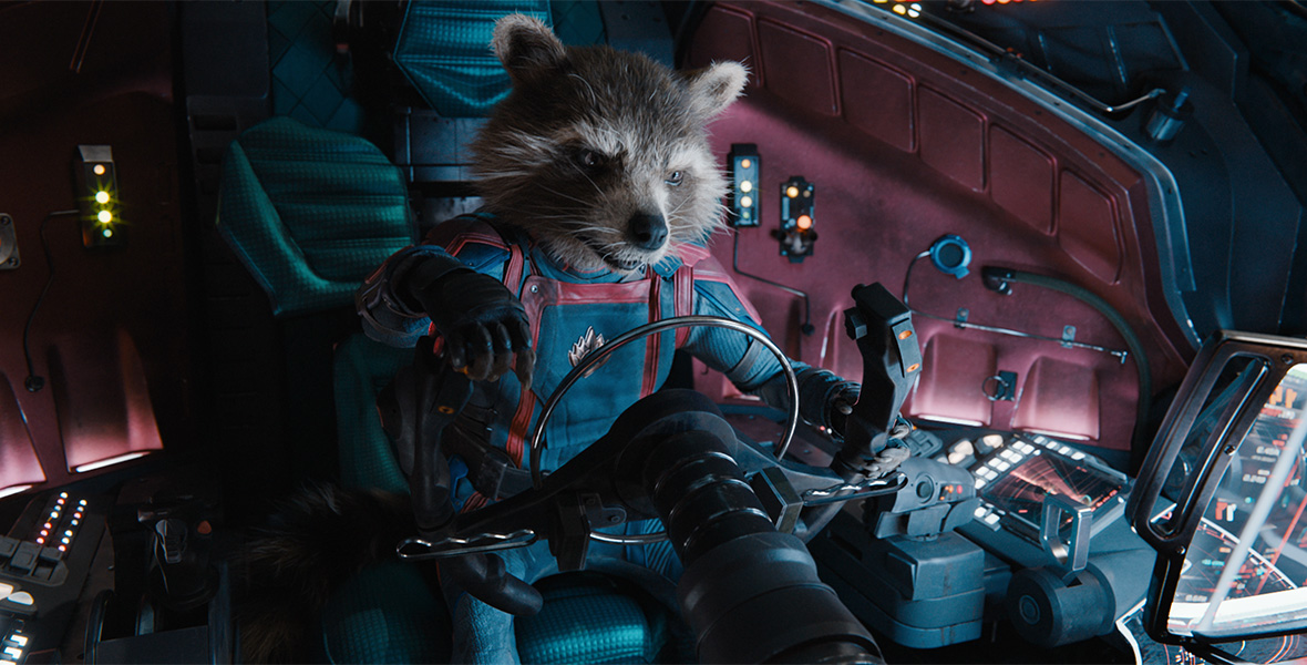 In a still from Marvel Studios’ Guardians of the Galaxy Vol. 3, Rocket (voiced by Bradley Cooper) is sitting behind a wheel and other controls on a spaceship, looking forward intently.