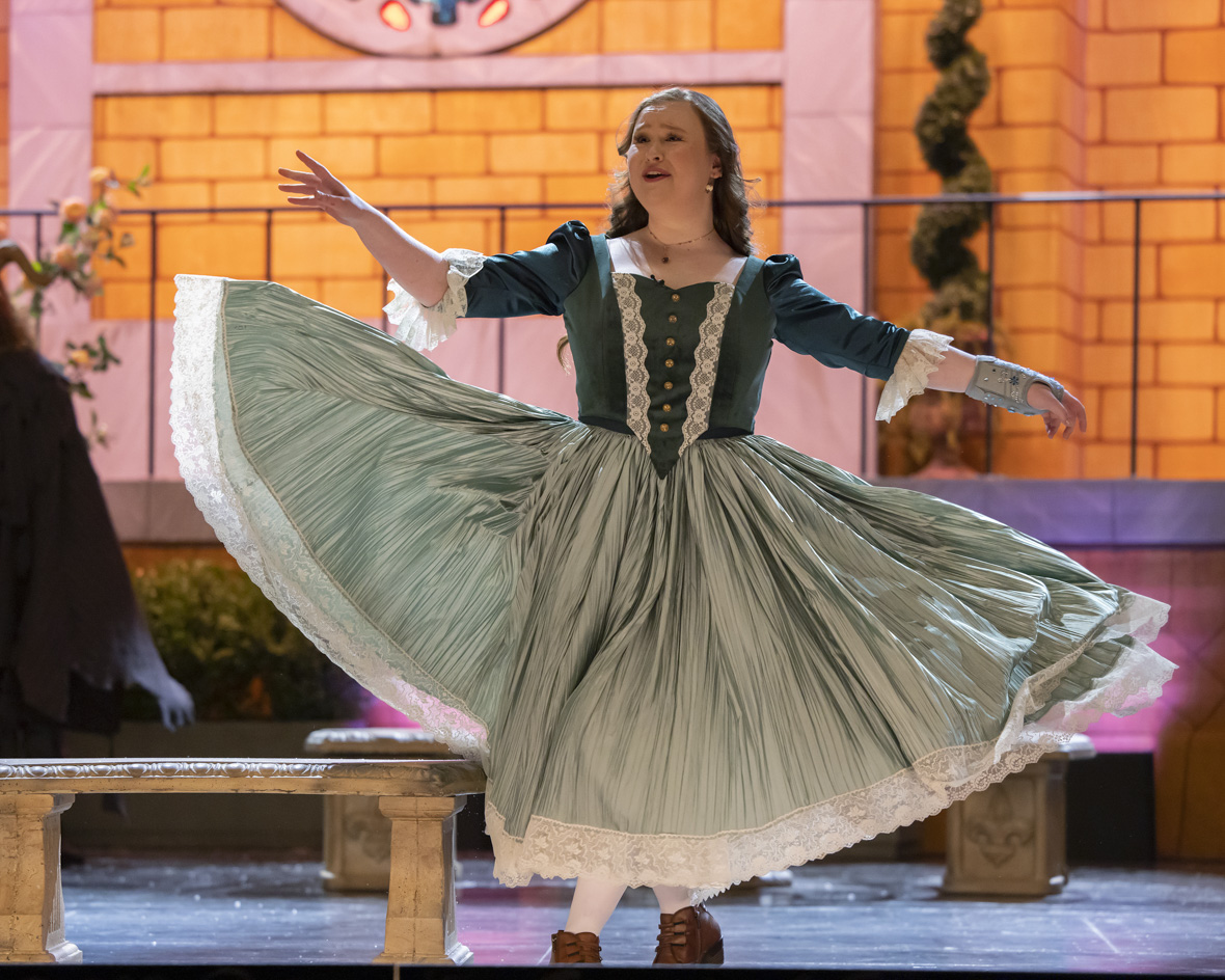Julia Lester as Ashlynn in High School Musical: The Musical: The Series. This photo is from a scene where Ashlynn is portraying Belle in Beauty and the Beast. Julia is onstage in a blue peasant dress and short brown boots. She has a brace around her left wrist and hand, indicating the character has an injury. The background behind her shows the exterior walls of a castle with some plants and trees. 