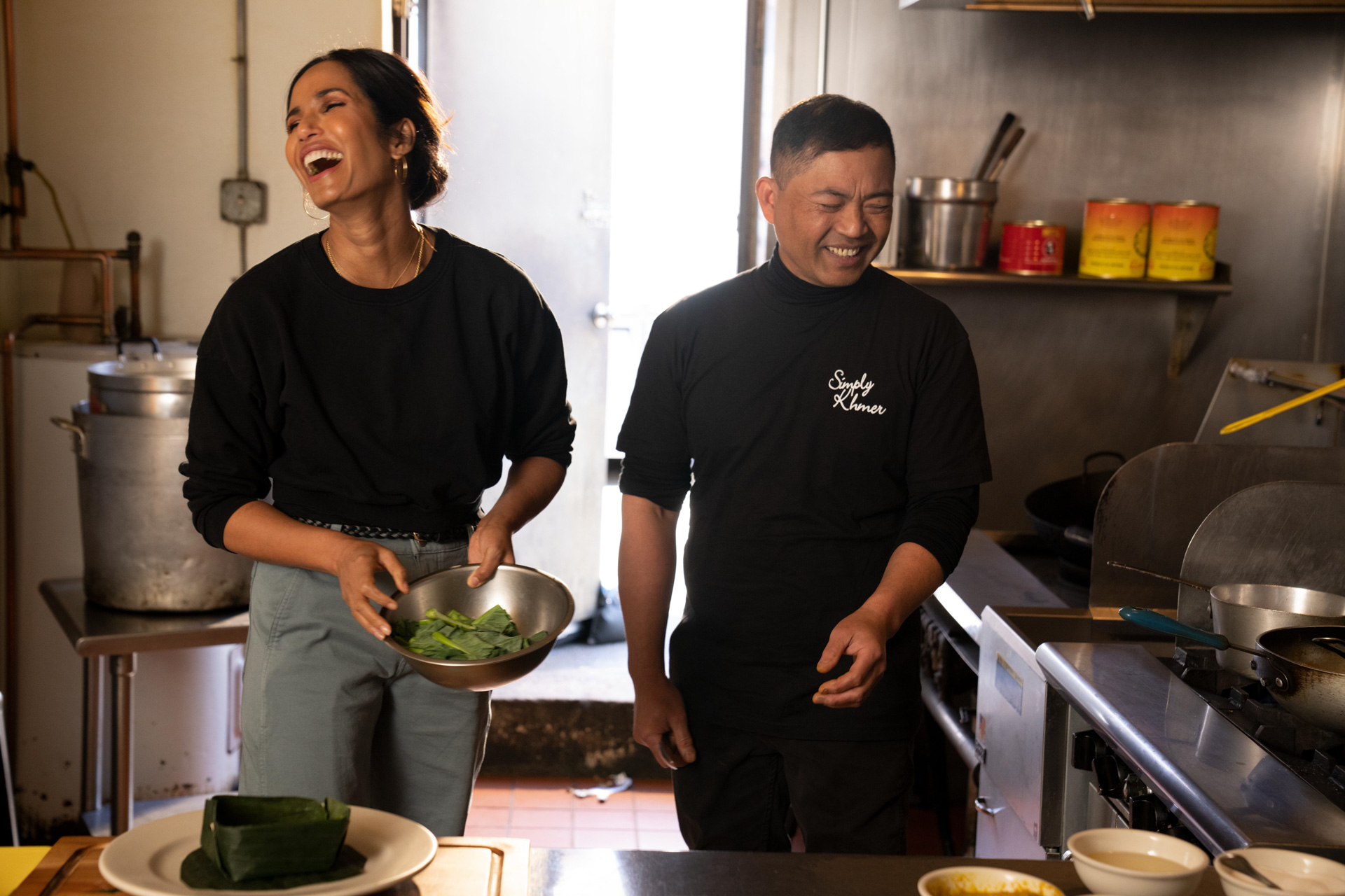 In a scene from Taste the Nation with Padma Lakshmi, Padma Lakshmi cooks in a restaurant and shakes a large silver bowl filled with greens.