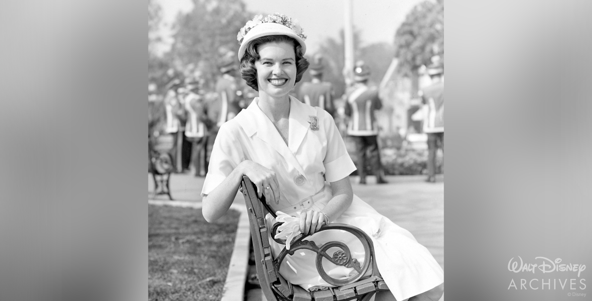 Valerie Curry, wearing a hat with flowers and a dress, sits on a bench at Disneyland Park and smiles at the camera. Behind her, a marching band is playing, and there are some trees in the distance.