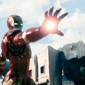In a scene from Iron Man, Iron Man wears a red and gold metal suit and fires a repulsor blast from his hand. A man cowers behind him while others put their hands up.