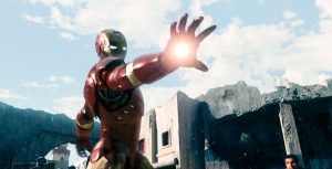 In a scene from Iron Man, Iron Man wears a red and gold metal suit and fires a repulsor blast from his hand. A man cowers behind him while others put their hands up.