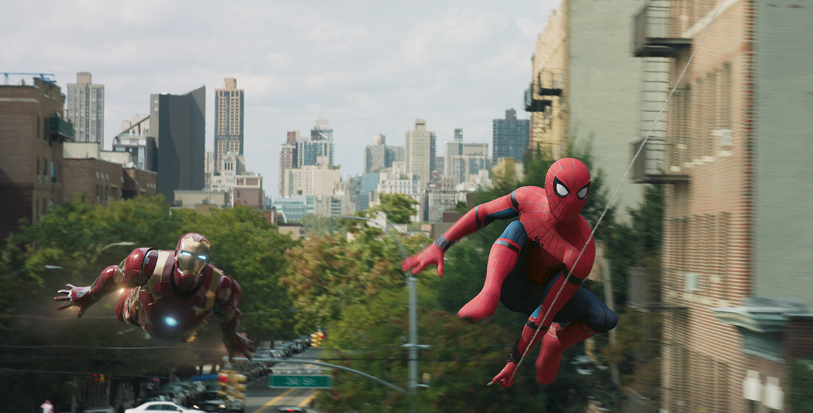 In a scene from Spider-Man: Homecoming, Iron Man (left) flies through a bustling New York City street as Spider-Man (right) swings from a web in front of him.